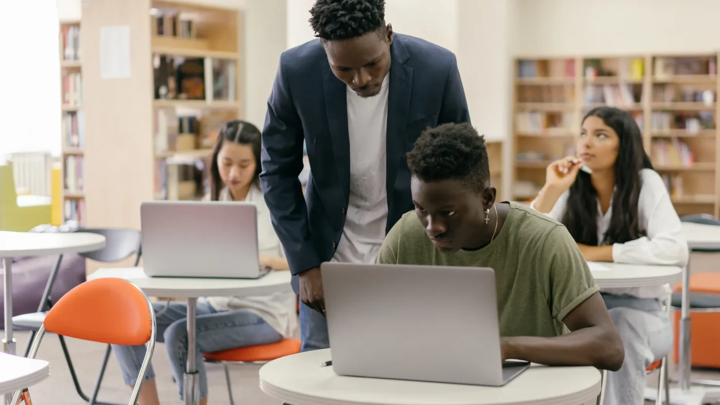 Students learning in a classroom setting with a teacher assisting and laptops on desks, creating an interactive education environment.
